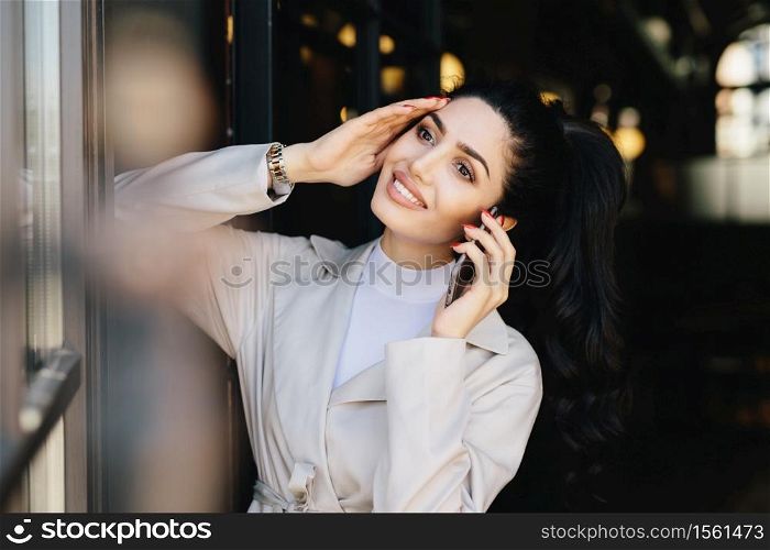Pretty brunette woman with delicate hands, alluring appearance communicating over cell phone while standing indoors. Young brunette businesswoman having rest speaking with her boyfriend using mobile