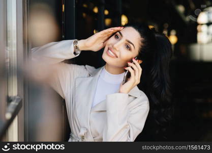 Pretty brunette woman with delicate hands, alluring appearance communicating over cell phone while standing indoors. Young brunette businesswoman having rest speaking with her boyfriend using mobile