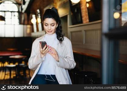 Pretty brunette woman with pony tail and appealing appearance sitting in cafe using her smartphone surfing internet using free wifi conncection and listening to her favourite music with earphones