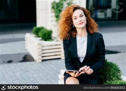 Pretty businesswoman with curly hair, pure skin and red lips, wearing formal costume while sitting at outdoor cafe or terrace, touching her modern table, communicating with friends using free wi-fi