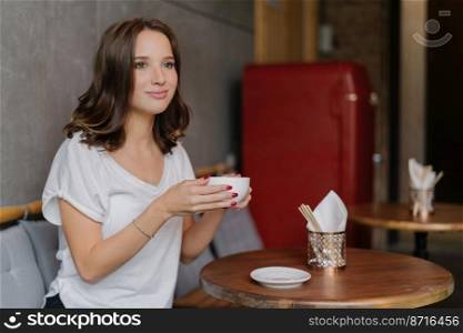Pretty carefree young woman in casual white t shirt, has happy face, drinks aromatic coffee, spends free time in restaurant, sits at round wooden table, has thoughtful expression into distance.