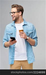 pretty casual man in blue jeans shirt holding his phone and cup of coffee to go standing over studio grey background.. pretty casual man in blue jeans shirt holding his phone and cup of coffee to go standing over studio grey background