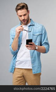 pretty casual man in blue jeans shirt holding his phone standing over studio grey background.. pretty casual man in blue jeans shirt holding his phone standing over studio grey background