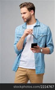 pretty casual man in blue jeans shirt holding his phone standing over studio grey background.. pretty casual man in blue jeans shirt holding his phone standing over studio grey background