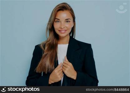 Pretty caucasian businesswoman with pleased face expression, charming smile, dressed in stylish black formal suit, sincerely gazing at camera on light blue background. Succesful women concept. Pretty businesswoman in formal suit on light blue background