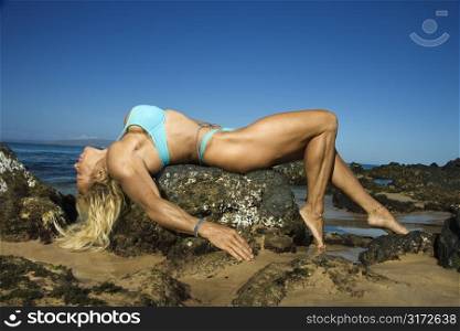 Pretty Caucasian mid adult woman bodybuilder in bikini lying on rock on Maui beach.