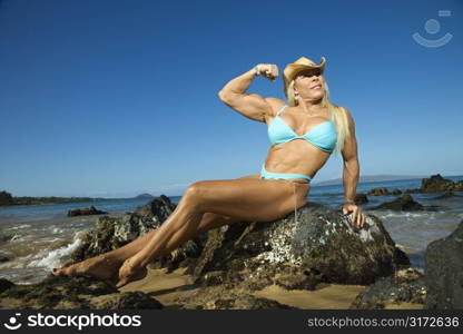 Pretty Caucasian mid adult woman bodybuilder in bikini sitting on rock flexing bicep on Maui beach.