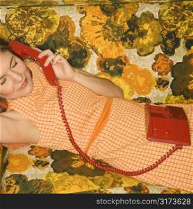 Pretty Caucasian mid-adult woman lying on colorful retro sofa talking on red telephone.