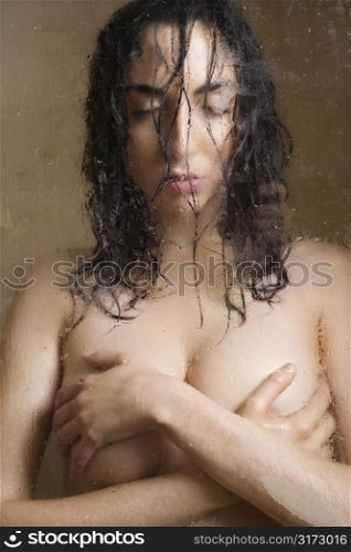 Pretty Caucasian nude young woman wet leaning against glass with arms over breasts.