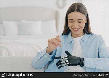 Pretty caucasian woman with artificial arm in bedroom. Happy girl is setting her electronic bionic limb. Smiling woman has modern cyber prosthesis. Engineering and technology concept.. Pretty caucasian woman with artificial arm in bedroom. Engineering and technology concept.