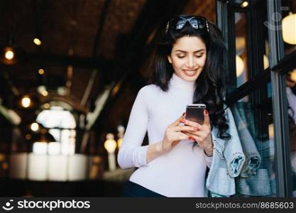 Pretty Caucasian woman with dark wavy hair wearing sunglasses and white elegant clothes holding smartphone using wireless Internet connection while chatting with her friends. Youth, lifestyle concept