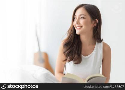 Pretty Charming Young Woman sitting smiling looking out at bedroom in the morning. Relaxed Beautiful female enjoying at home. Cheerful Asian girl reading book in white room. Healthy Lifestyle, Holiday