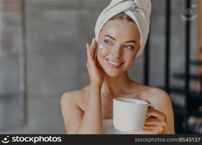 Pretty charming young woman touches face gently, applies face cream, wears minimal makeup, smiles gently, wears wrapped white bath towel on head, drinks aromatic coffee, spends free time at home
