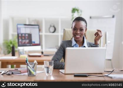 Pretty cheerful african businesswoman working with laptop in office and looking at camera