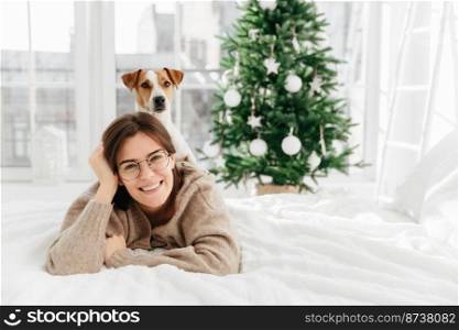 Pretty cheerful woman wears big optical round glasses, lies on bed, her dog poses on back, have fun together, spend winter holidays at home, decorated green Christmas tree. Coziness, winter, festivity