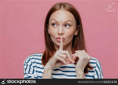 Pretty curious Caucasian woman makes silence sign, keeps index finger over mouth, looks secretly aside, wears striped casual clothes, poses over pink studio wall with free space for your text. Hush