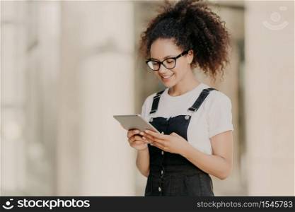 Pretty curly haired girl looks at digital tablet screen with smile, reads some text or electronic book, wears spectacles for vision correction, white t shirt and dungarees, enjoys high speed internet