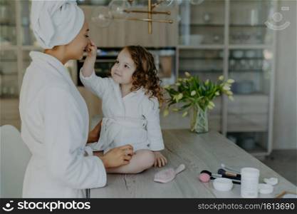 Pretty curly small girl touches nose of her mother dressed in white soft dressing gown sits on table with cosmetic products going to do makeup for mom pose together against cozy home interior