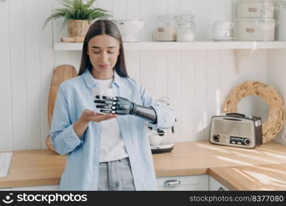 Pretty disabled girl is holding cup of tea with cyber hand carefully. Concept of grasp sensors in modern bionic prosthesis. Routine of handicapped person and life quality. Woman at domestic kitchen.. Pretty disabled girl is holding cup of tea with cyber hand carefully. Concept of grasp sensors.