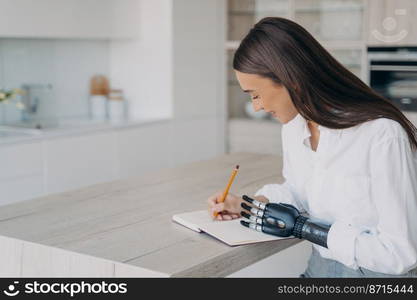 Pretty disabled girl with bionic prosthetic arm writing in notebook, sitting at kitchen table at home. Young woman with disability makes notes, planning day, using artificial robotic hand.. Pretty disabled girl with bionic prosthetic arm writing in notebook sitting at kitchen table at home