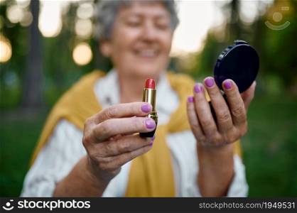 Pretty elderly woman holds lipstick in summer park, back to youth. Aged people lifestyle. Funny grandmother having fun outdoors, old female person on nature. Pretty elderly woman holds lipstick in summer park