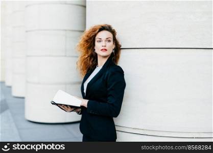 Pretty elegant woman with curly wavy hair dressed in black suit, holding pocket book in hands looking aside into distance, thinking over her future plans. Fashionable lady writing necessary notes