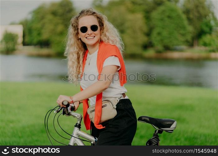 Pretty female bike rider stops near beautiful river, wears sunglasses, white t shirt, jumper on shoulders, poses outdoor with her transport, expresses positive emotions, enjoys awesome travel