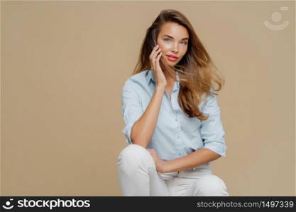 Pretty female feels relaxed while sits at chair, dressed in stylish shirt and white trousers, poses over brown hair, copy space for your promotional content. Beautiful woman in fashionable clothes