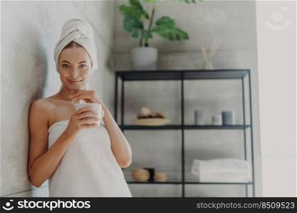 Pretty female model has perfect skin, touches jawline gently, drinks hot beverage, feels refreshed and relaxed, wrapped in bath towel, poses in bathroom. Natural beauty and skin care concept