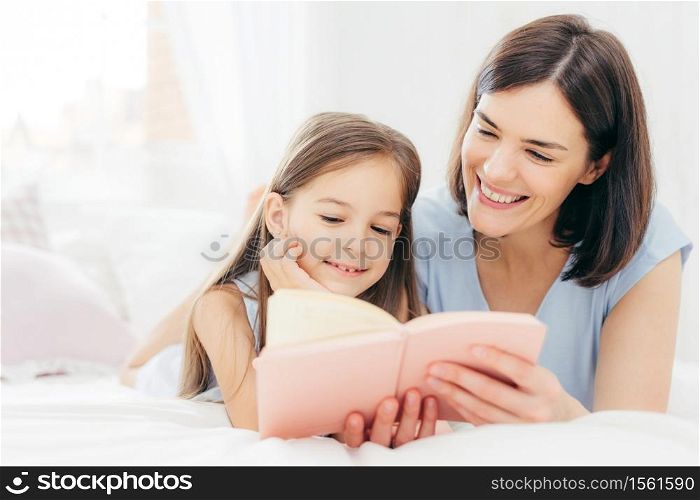 Pretty female mother and her small daughter read interesting book in bedroom, lie on comfortabled bed during morning. Lovely mum reads fairty tale for her little female kid, who is very curious