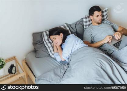 Pretty female sleeps in bed, sees pleasant dreams, while her husband works on laptop computer, prepares report, tries not to interrupt his wife sleep. Family couple in bedroom. Rest concept.