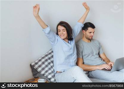 Pretty female stretches as just awoke, has good morning, enjoys day off and her husband sits near in bed, plays computer games on laptop, being concentrated. Family, leisure and home concept
