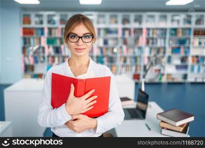 Pretty female student in glasses with opened book in university library. Young woman in reading room, knowledge depository