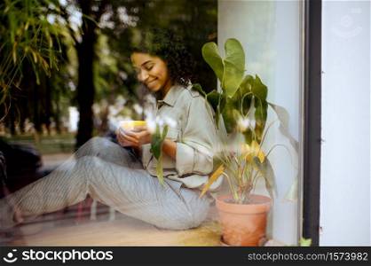 Pretty female student with cup of coffee sitting on the windowsill in cafe. Woman learning a subject in coffeehouse, education and food. Girl studying in campus cafeteria. Student with coffee sitting on windowsill in cafe