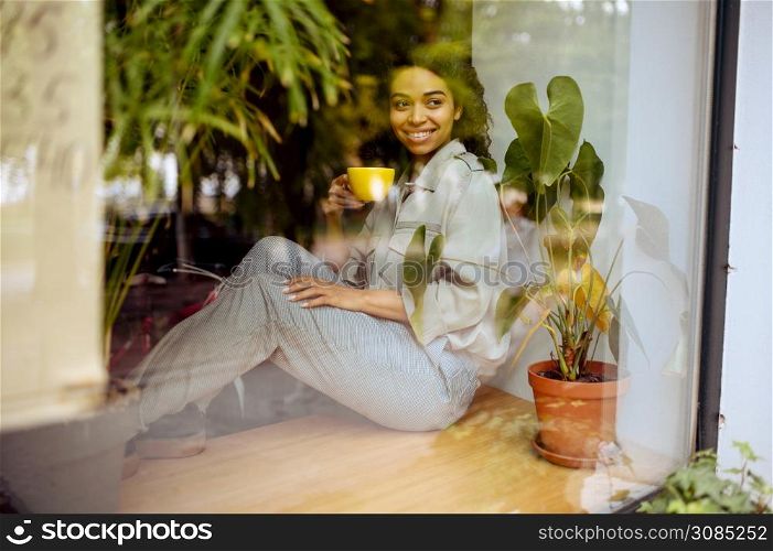 Pretty female student with cup of coffee sitting on the windowsill in cafe. Woman learning a subject in coffeehouse, education and food. Girl studying in campus cafeteria. Student with coffee sitting on windowsill in cafe