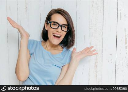 Pretty female with dark hair, pleasant appearance, looks joyfully at camera, keeps palms raised, gestures happily, wears casual t shirt and spectacles, stands against white wooden background