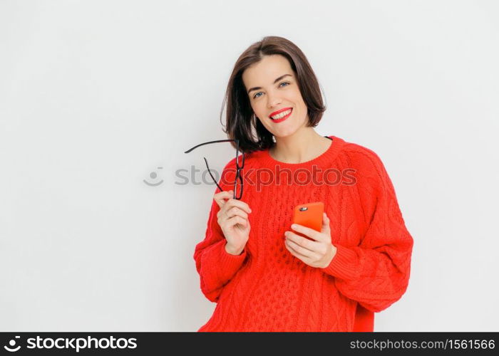 Pretty female with dark short hair, wears oversized red winter sweater, holds eyewear and smart phone, happy to message with friends, stands against white background. Happiness and technology concept