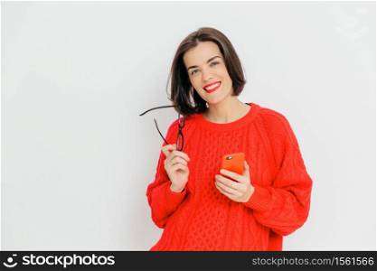Pretty female with dark short hair, wears oversized red winter sweater, holds eyewear and smart phone, happy to message with friends, stands against white background. Happiness and technology concept