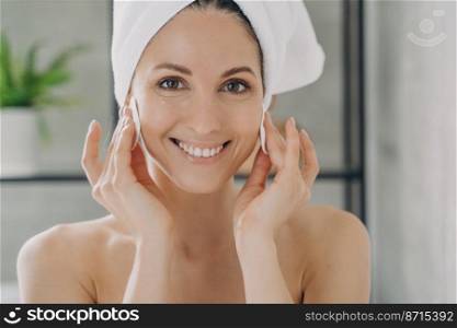 Pretty female with towel on head cleansing face, removing makeup, using soft cotton pads. Smiling hispanic woman wiping cheeks with moisturizing lotion or skin cleanser. Beauty treatment, skincare.. Happy woman cleansing face, removing makeup by cotton pads after shower. Beauty treatment, skincare