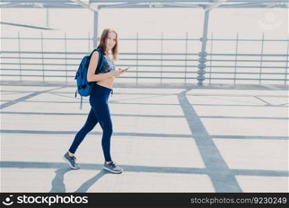 Pretty female youngster has walk outside, carries rucksack, holds modern cell phone, listens music, checks favourite song in playlist, wears casual sportswear and sneakers, has healthy lifestyle