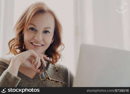 Pretty ginger businesswoman searches online web informationn via laptop computer, has pleasant smile, holds optical glasses, sits at coworking desktop, busy with freelance work. Technology concept