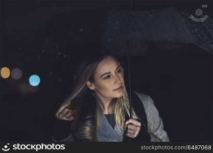 Pretty girl at rainy night outdoors, portrait of a beautiful blond female standing with umbrella under rainfall, good mood in cold wet weather. Pretty girl at rainy night outdoors