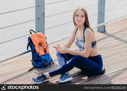 Pretty girl has satisfied expression after going in for sport, enjoys favourite song in earphones, holds cellular, messages with friends, poses crossed legs. Technology and lifestyle concept