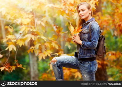 Pretty girl in autumn park, nice female standing with dry maple leaves in hands and enjoying last warm sunny autumnal days