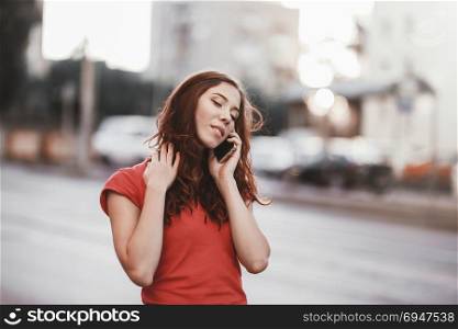 Pretty girl in casual dress is talking on a mobile phone while walking at the city street on a sunset time.. Pretty girl in casual dress is talking on a mobile phone while walking at the city street on a sunset time