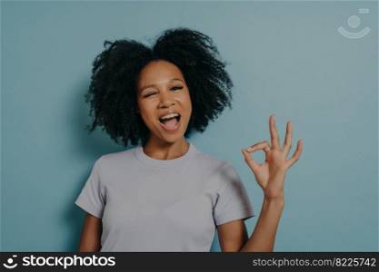 Pretty girl of mixed race appearance showing ok gesture with fingers and winks, showing that everything is great. Attractive African female in good mood posing in studio, looking friendly at camera. Pretty girl of mixed race appearance showing ok gesture with fingers and winks