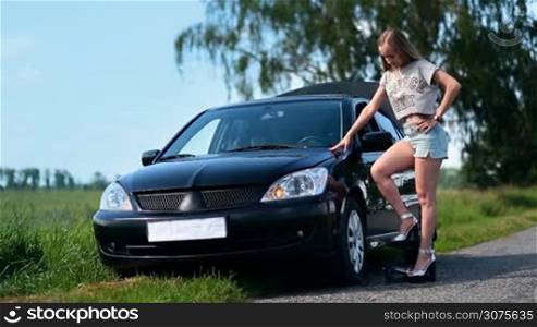 Pretty girl on high heels is inflating car tyre with foot pump on country road
