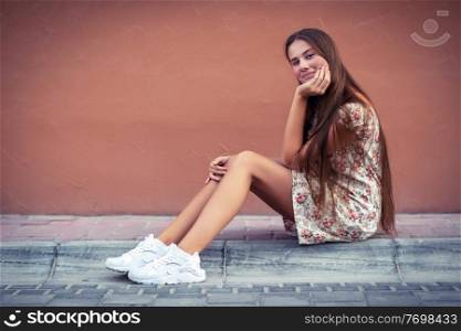 Pretty girl on the street, nice female with long natural hair sitting on blank monophonic wall background, authentic genuine beauty of young people
