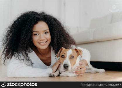 Pretty girl with Afro hair, lies on floor with dog, expresses pleasant emotions, poses in living room near couch, bought pet in new apartment. Woman host with beloved animal at home, share good moment