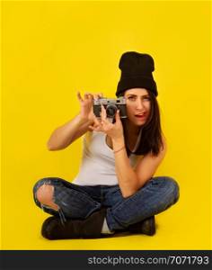 Pretty girl with dark hair in casual hipster clothes and retro camera sitting cross-legged on a bright yellow background
